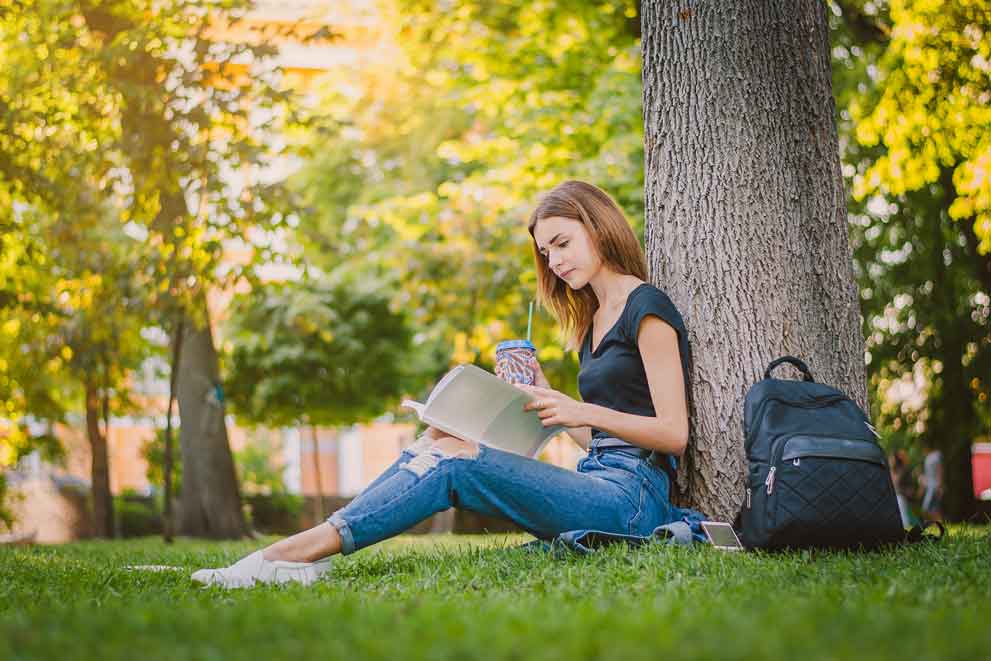 A female summer school student reading a book outside under a tree in the sunshine